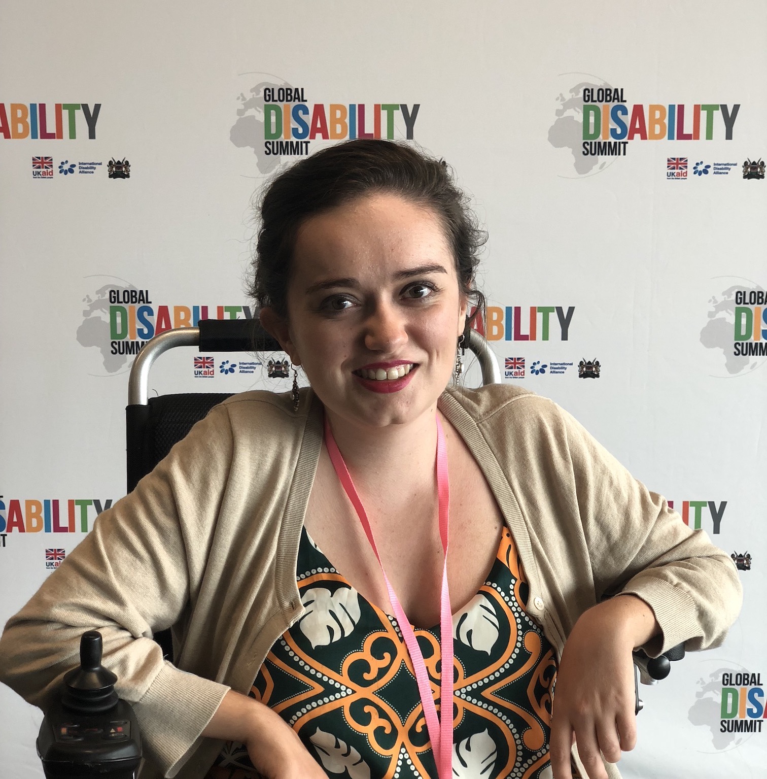 A white woman with brown hair pulled back, in a green patterned dress and beige cardigan, sitting in a wheelchair, smiling like she loves having a headshot taken